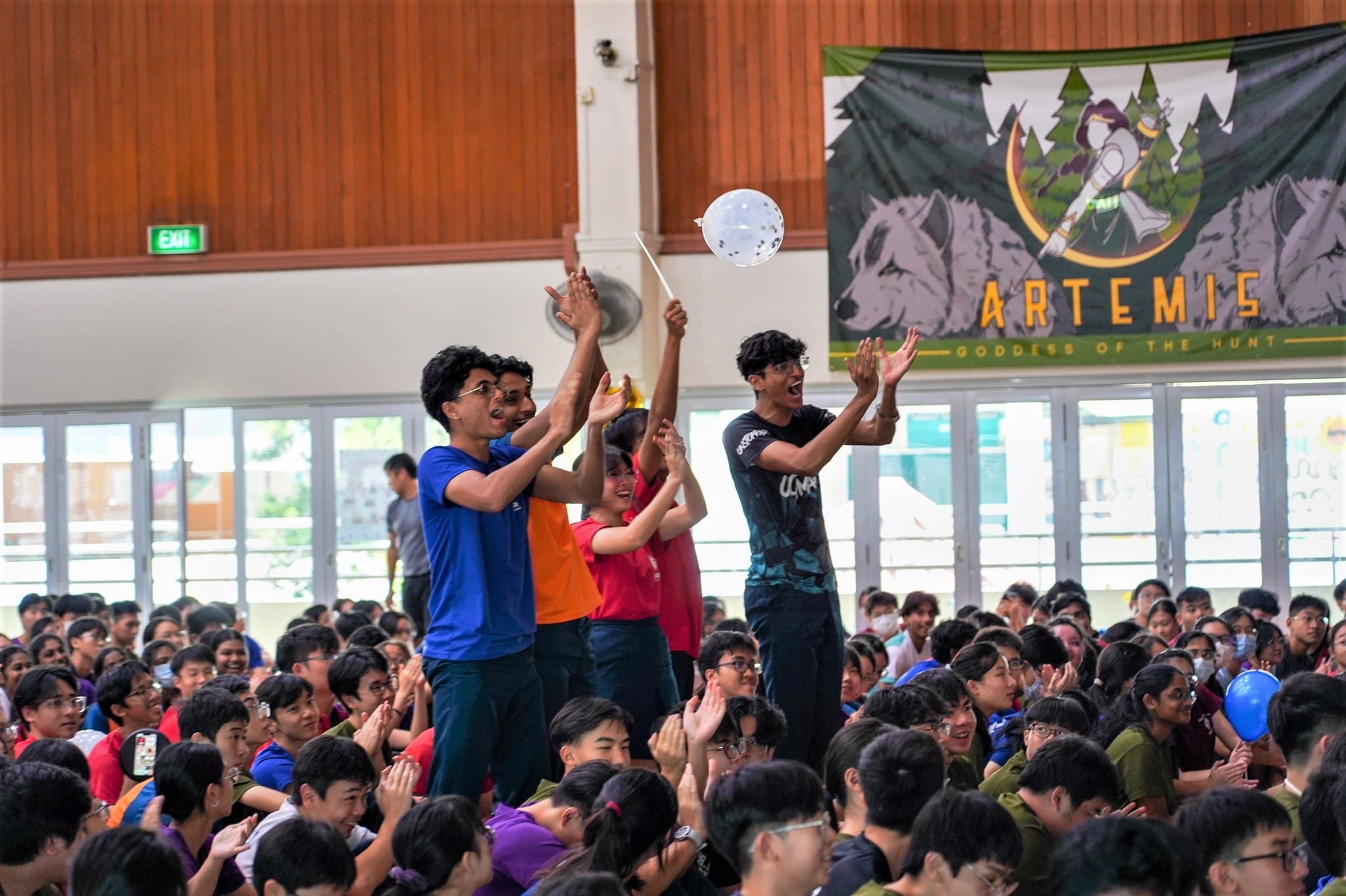 Students cheer in a crowded hall with an Artemis Goddess of the Hunt banner, as one holds a balloon on a stick.